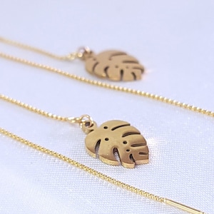 May include: A pair of gold-tone dangle earrings with a monstera leaf design. The earrings are made of a thin chain with a small leaf charm at the end.