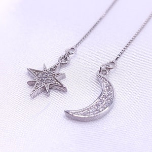 以下が含まれることがあります： 星と三日月のチャームが付いたシルバーのドロップイヤリングのペア。各チャームには、小さなクリアクリスタルが埋め込まれています。