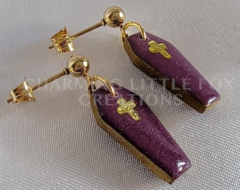 May include: A pair of purple coffin-shaped earrings with gold accents and a small gold cross on each coffin. The earrings are on a white background.