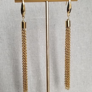 May include: A pair of gold-colored tassel earrings. Each earring features a teardrop-shaped top connected to a cylindrical piece, from which a long chain tassel hangs. The earrings are displayed on a gold-colored jewelry stand against a neutral background.