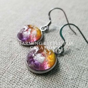 Könnte beinhalten: Ein Paar silberne Ohrhänger mit runden, bunten Harz-Cabochons. Die Cabochons sind eine Mischung aus Lila, Weiß und Gelb.