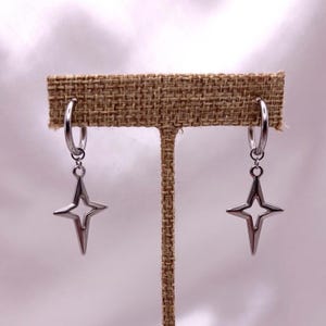 May include: A pair of silver-colored hoop earrings with a starburst charm. The earrings are displayed on a brown, textured surface. The starburst charm has a four-pointed star design.