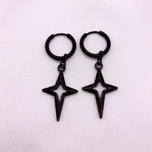 May include: A pair of black hoop earrings with a starburst design. Each earring features a small hoop that connects to a star-shaped pendant with a cut-out center. The earrings are displayed against a white background.