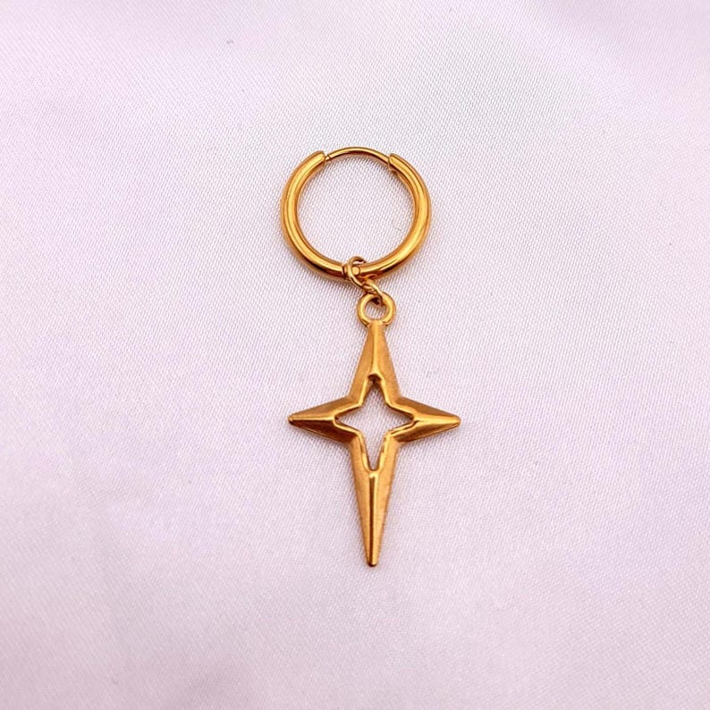 May include: A gold-colored earring featuring a star pendant. The star has a four-pointed design with a cutout center. The earring is suspended from a circular hoop. The jewelry is set against a white background.