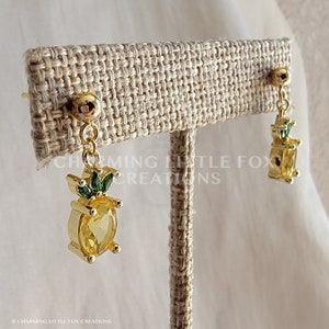 May include: Gold pineapple earrings with green crystal accents. The earrings are on a brown burlap display stand.