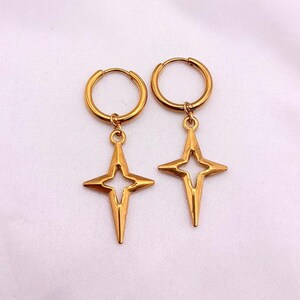 May include: A pair of gold-colored hoop earrings with a starburst pendant. The earrings have a simple, elegant design, with a circular hoop and a four-pointed star charm. The earrings are displayed on a white background.