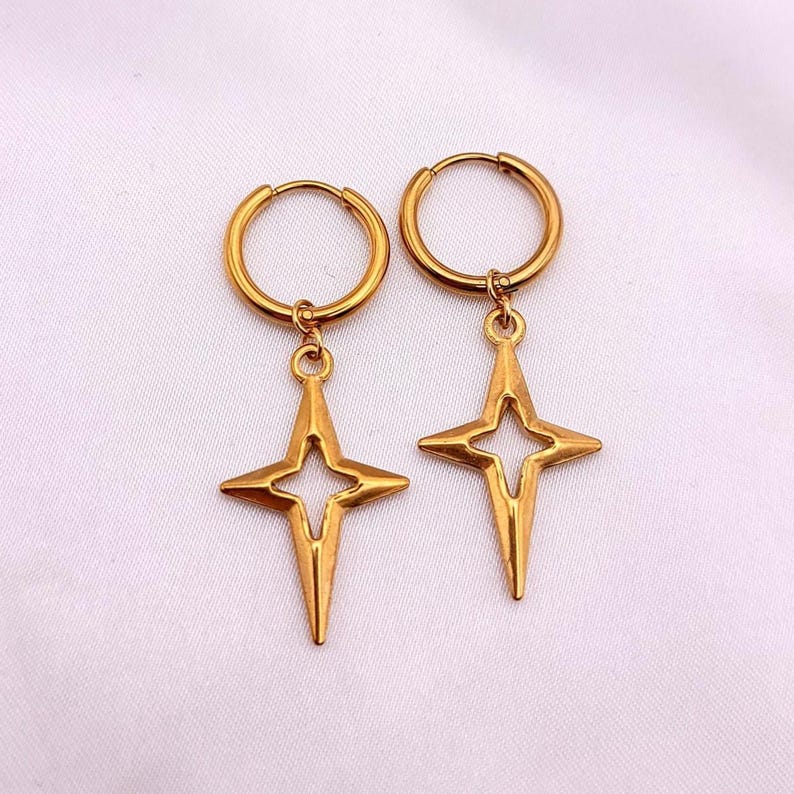 May include: A pair of gold-colored hoop earrings with a starburst pendant. The earrings have a simple, elegant design, with a circular hoop and a four-pointed star charm. The earrings are displayed on a white background.