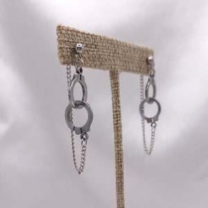 May include: A pair of silver handcuff earrings with a chain dangle. The earrings are displayed on a brown burlap earring stand.