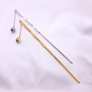 Könnte beinhalten: Eine silberfarbene Metall-Haarnadel und eine goldfarbene Metall-Haarnadel, beide mit gewelltem Schaft. Jede Haarnadel hat eine kleine Kette am oberen Ende, an der ein kleiner, polyedrischer d20-Spielwürfel als Anhänger befestigt ist.