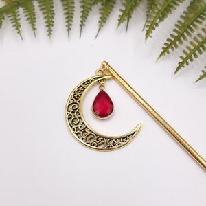 May include: Gold hair stick with a crescent moon design and a dangling red teardrop-shaped crystal.