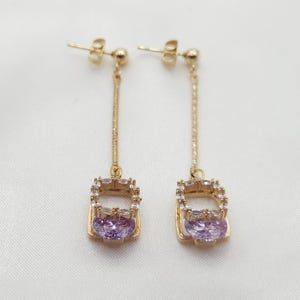 Puede incluir: Un par de pendientes colgantes de oro con un colgante de forma cuadrada que presenta una piedra preciosa p&uacute;rpura rodeada de diamantes de imitaci&oacute;n transparentes.
