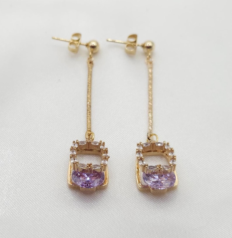 Puede incluir: Un par de pendientes colgantes de oro con un colgante de forma cuadrada que presenta una piedra preciosa p&uacute;rpura rodeada de diamantes de imitaci&oacute;n transparentes.
