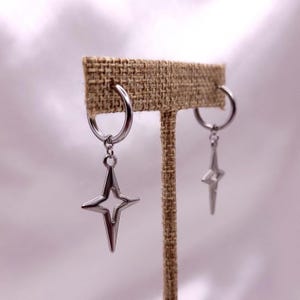 May include: Silver-colored hoop earrings with a four-pointed star pendant. The earrings are displayed on a tan fabric earring holder. The star pendant has a cut-out center and a polished finish, reflecting light.