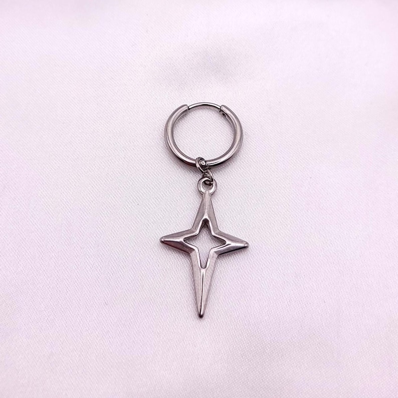 May include: A single silver-colored earring with a star-shaped pendant. The earring features a hoop design with a star pendant hanging from it. The star has a four-pointed design with a cutout in the center. The earring is displayed against a white background.