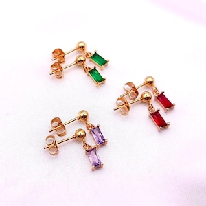May include: Gold-colored stud earrings with a small, round stud and a dangling rectangular gemstone. The earrings are available in green, red, and purple. The earrings are displayed on a white surface.