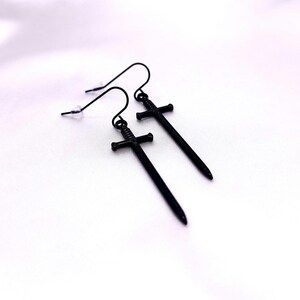 May include: A pair of black sword earrings with a simple design. The swords have a cross-shaped guard and a pointed tip.