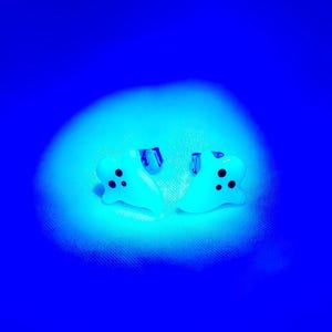 May include: A pair of ghost-shaped earrings. Each earring is white with three black dots for eyes and a mouth. The earrings are set against a blue background with a bright, circular light.