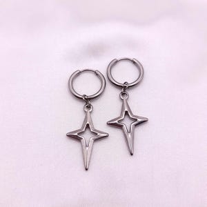 May include: A pair of silver-colored hoop earrings. Each earring features a star-shaped pendant with a four-pointed design. The earrings are displayed against a soft, white background.