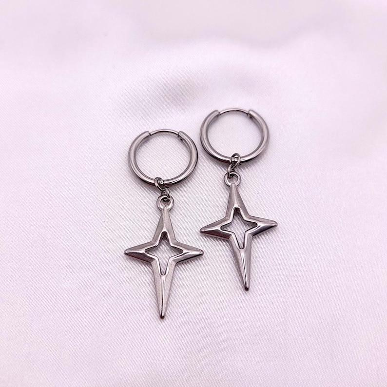 May include: A pair of silver-colored hoop earrings. Each earring features a star-shaped pendant with a four-pointed design. The earrings are displayed against a soft, white background.