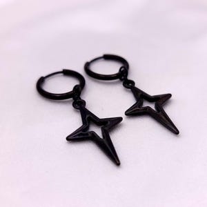 May include: A pair of black hoop earrings with star-shaped pendants. The earrings are made of a dark metal and feature a simple, geometric design. The star pendants have a pointed, open-center design.