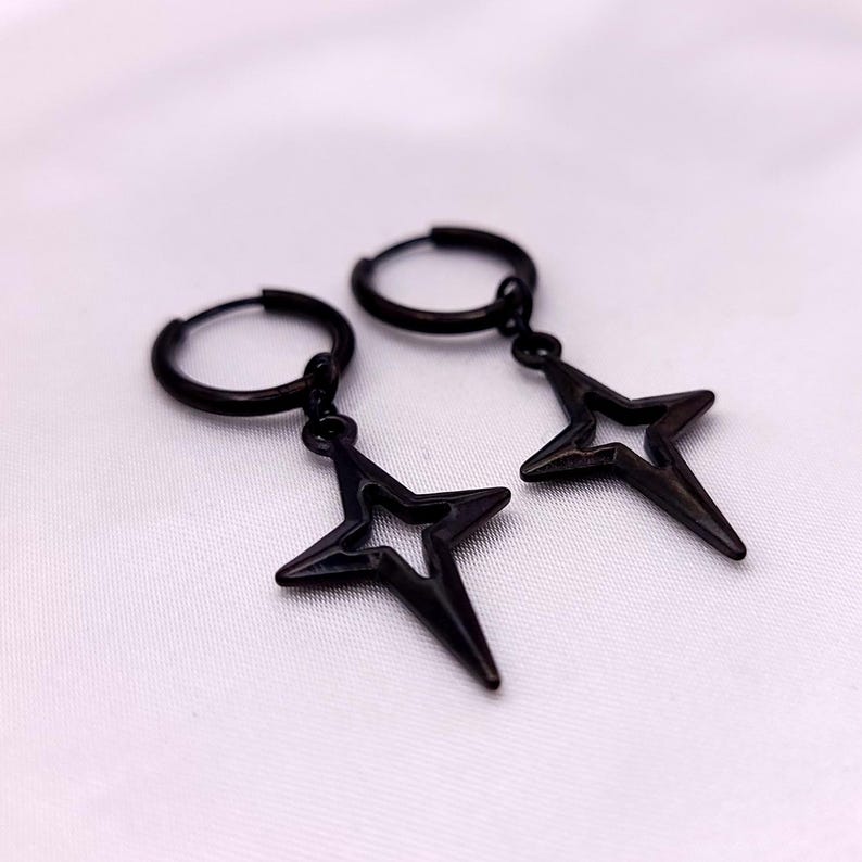 May include: A pair of black hoop earrings with star-shaped pendants. The earrings are made of a dark metal and feature a simple, geometric design. The star pendants have a pointed, open-center design.