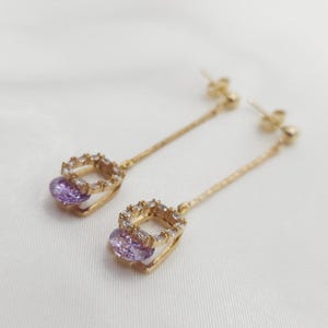 Puede incluir: Un par de pendientes colgantes de oro con una piedra preciosa p&uacute;rpura y detalles de cristal transparente.