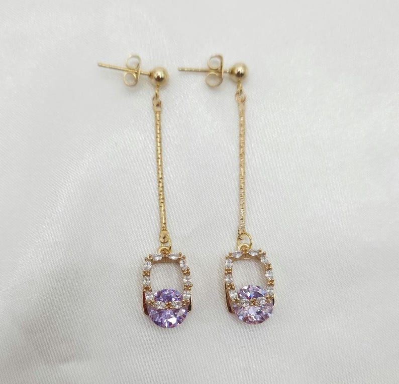 Puede incluir: Un par de pendientes colgantes de oro con un colgante de forma cuadrada que presenta una piedra preciosa p&uacute;rpura rodeada de piedras transparentes m&aacute;s peque&ntilde;as.