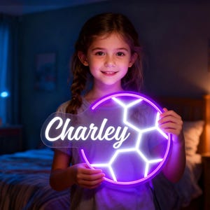 May include: A neon sign in the shape of a soccer ball with the name "Charley" in white script. The sign has a purple outline and white interior, held by a smiling person. The background is a softly lit room.