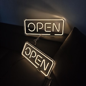 以下が含まれることがあります： 2つの「OPEN」ネオンサイン。各サインは、丸みを帯びた角と白いネオンの輝きを持つ長方形の形状をしています。「OPEN」の文字は、太字のサンセリフ体で書かれています。サインは暗い背景にあります。