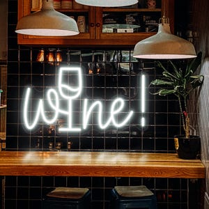 Peut inclure: Enseigne au néon blanche avec le mot "Wine!" et un symbole de verre à vin. Le signe est fixé sur un mur carrelé noir, avec un bar en bois et des tabourets. Deux lampes suspendues éclairent la scène.
