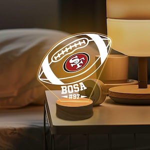 Può includere: Lampada da notte decorativa a forma di pallone da football con il logo dei 49ers di San Francisco. La lampada ha una base in legno e mostra il nome "BOSA" e il numero "97". Emette una luce calda.