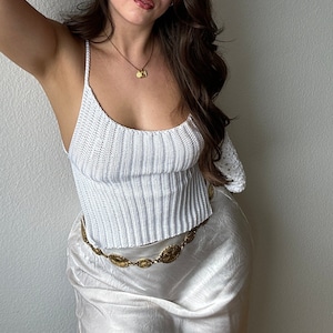 May include: A woman wearing a white crocheted top with a gold chain belt and a white satin skirt.