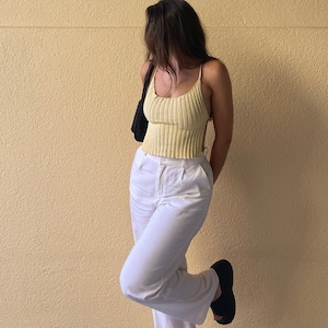 May include: A woman wearing a yellow crochet tank top and white pants. She is standing in front of a yellow wall.