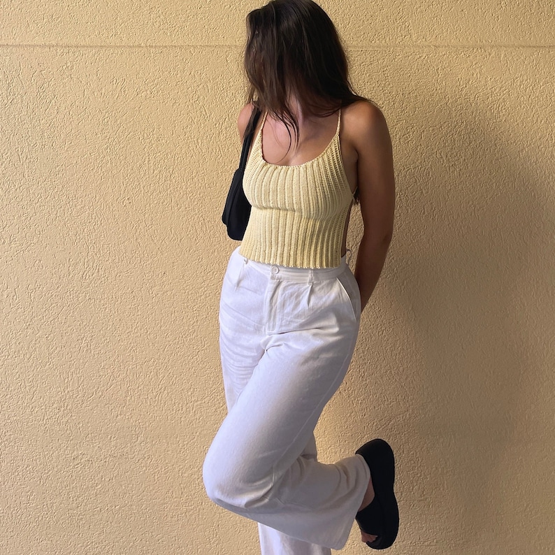 May include: A woman wearing a yellow crochet tank top and white pants. She is standing in front of a yellow wall.