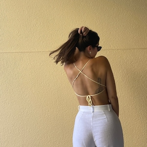 May include: A woman wearing a white halter top with a criss-cross back design. The top is made of a thin, stringy material. The woman is wearing white pants and has her hair pulled back in a ponytail.