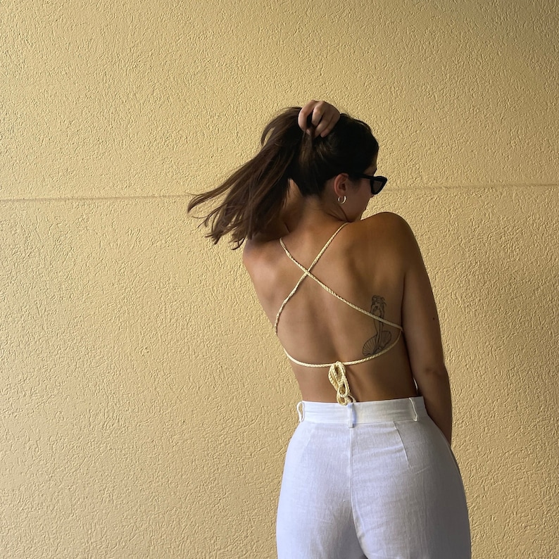 May include: A woman wearing a white halter top with a criss-cross back design. The top is made of a thin, stringy material. The woman is wearing white pants and has her hair pulled back in a ponytail.