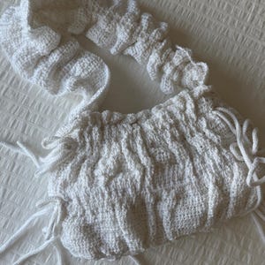 May include: A white crocheted shoulder bag featuring a gathered, textured surface with a ruffled strap. The bag includes side panels with decorative drawstring ties that hang loosely from the edges, creating a soft, dimensional appearance.
