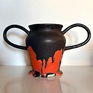 May include: A ceramic vessel featuring two large, curved handles on either side. The upper portion is finished in a matte black glaze, while the lower section displays a vibrant, dripping orange glaze that extends down to the base of the object.