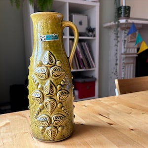 Puede incluir: Un jarrón de cerámica verde con un diseño floral y una etiqueta que dice "gastliches heim".