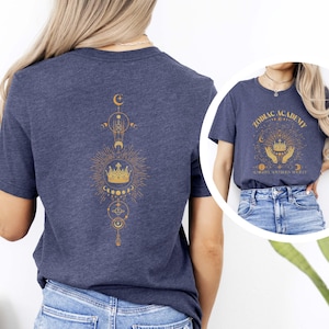 Pu&ograve; includere: Due t-shirt blu navy con disegni dorati a tema zodiacale. La parte anteriore di una t-shirt presenta il testo "Zodiac Academy" con una corona e mani che tengono la corona. La parte posteriore dell'altra t-shirt presenta una corona con un design di luna e stelle.