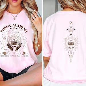 Pu&ograve; includere: Maglietta rosa con un design grafico di una corona, stelle e il testo "Zodiac Academy Almighty Sovereign Society". Il retro della maglietta ha un design simile con una corona e simboli celesti.