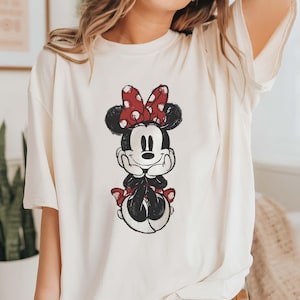 May include: A white t-shirt with a black and red cartoon image of Minnie Mouse. Minnie Mouse is wearing a red bow with white polka dots and a red skirt with white polka dots.