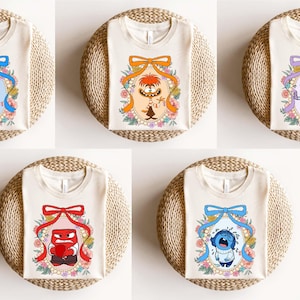 May include: Five cream-colored t-shirts, each featuring a different character from the animated movie "Inside Out." Each shirt has a unique character design framed by a floral border and a decorative bow.