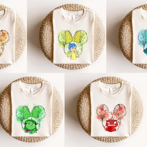 May include: Five white t-shirts with different watercolor illustrations of characters from the movie Inside Out. Each character is depicted with a Mickey Mouse ear design. The characters are Joy, Sadness, Anger, Disgust, and Fear.