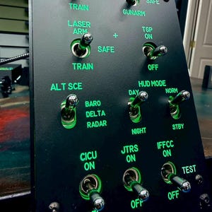 Peut inclure: Un panneau de contrôle noir avec plusieurs interrupteurs à bascule et du texte vert éclairé. Le texte comprend des étiquettes telles que "MASTER A", "TRAIN", "SAFE", "GUNARM", "LASER ARM", "TGP ON", "HUD MODE", et "IFFCC ON".