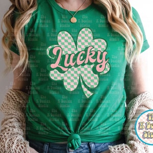 May include: Green t-shirt featuring a pink and green checkered shamrock design with the word "Lucky" in pink script. The shirt is a crew neck and has a knot at the bottom. The K Design Studio logo is visible.