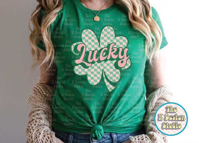 May include: Green t-shirt featuring a pink and green checkered shamrock design with the word "Lucky" in pink script. The shirt is a crew neck and has a knot at the bottom. The K Design Studio logo is visible.