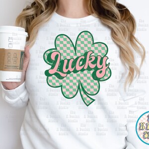 May include: White sweatshirt featuring a green and pink checkered shamrock with the word "Lucky" in a retro font. The design includes a green outline and a small circular logo with text. A coffee cup is held in the image.