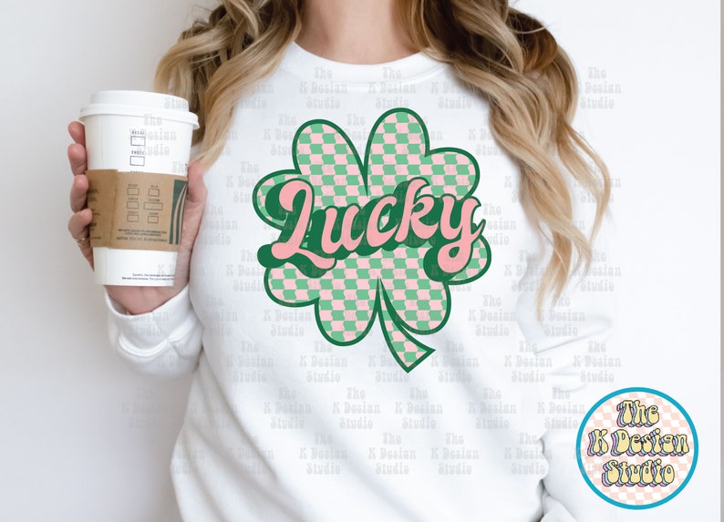 May include: White sweatshirt featuring a green and pink checkered shamrock with the word "Lucky" in a retro font. The design includes a green outline and a small circular logo with text. A coffee cup is held in the image.