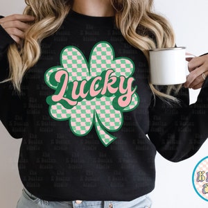 May include: Black sweatshirt featuring a large, retro-style shamrock graphic with a checkered pattern in green and pink. The word "Lucky" is written across the shamrock in a cursive font. A white mug is held in the image.